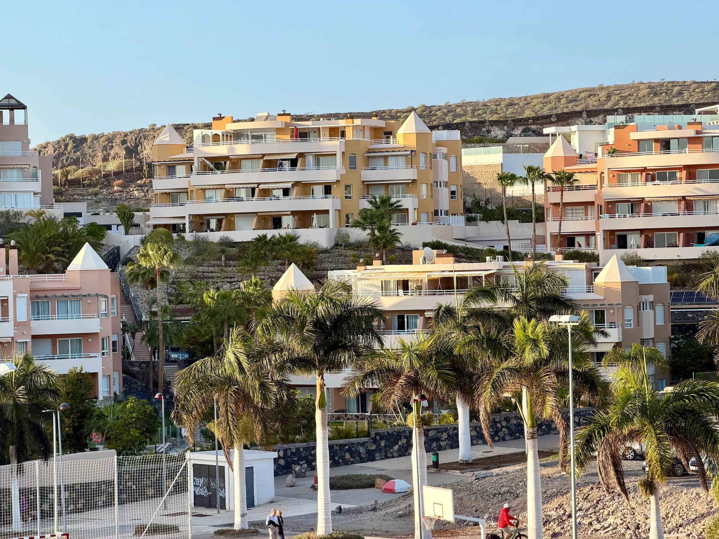 Kaskadowe, pomarańczowe budynki mieszkalne na wzgórzu z palmami i boiskiem sportowym na pierwszym planie