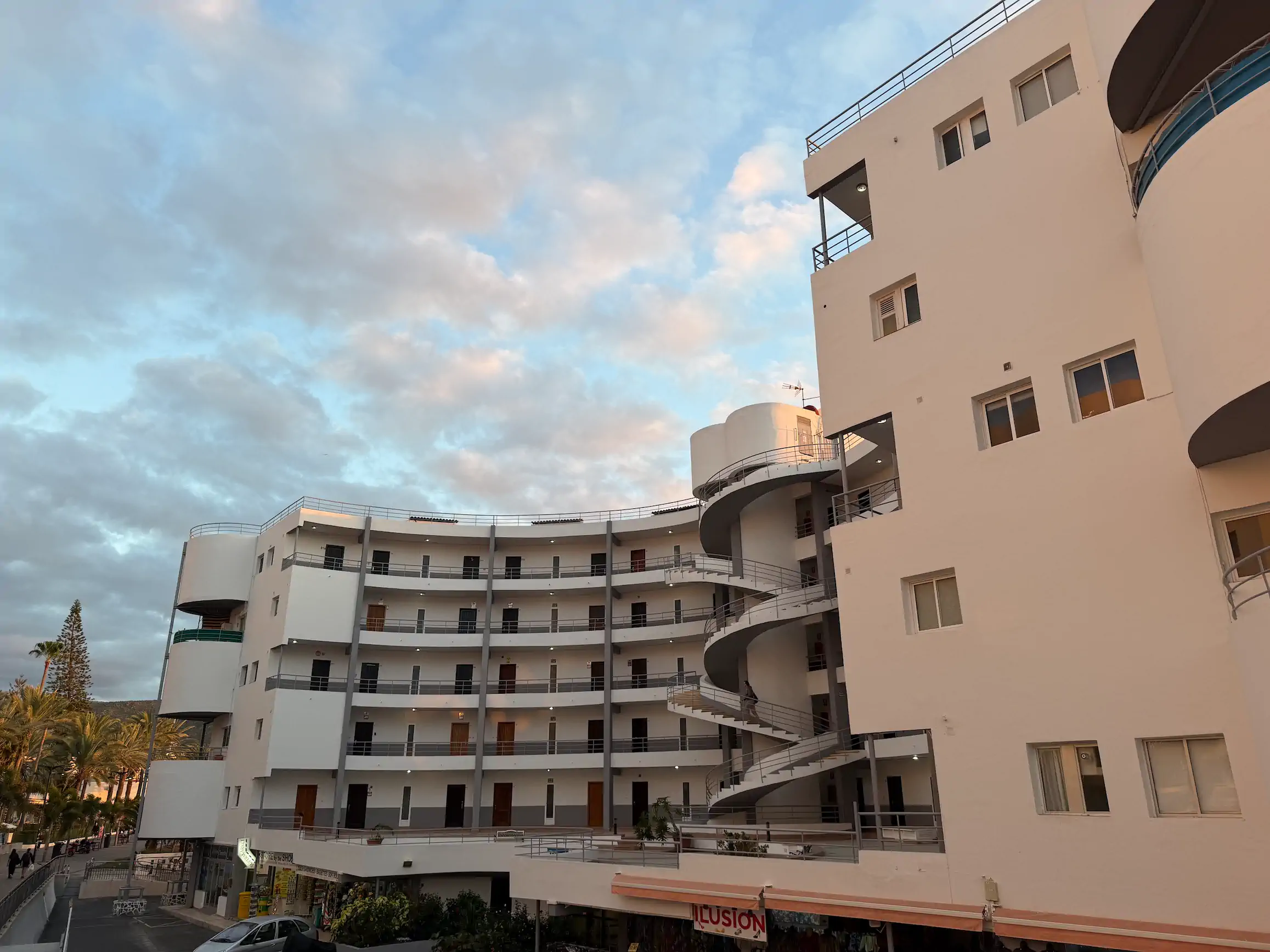 Biały budynek apartamentowy o zakrzywionym kształcie z zewnętrznymi korytarzami i spiralną klatką schodową w świetle zachodzącego słońca