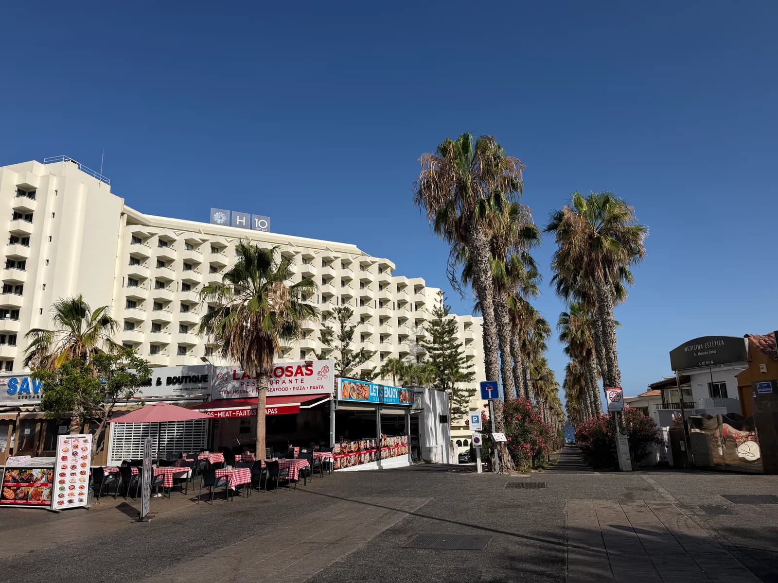 Biały budynek hotelowy, restauracje i wysokie palmy przy Avenida Rafael Puig Lluvina w Playa de las Américas o poranku