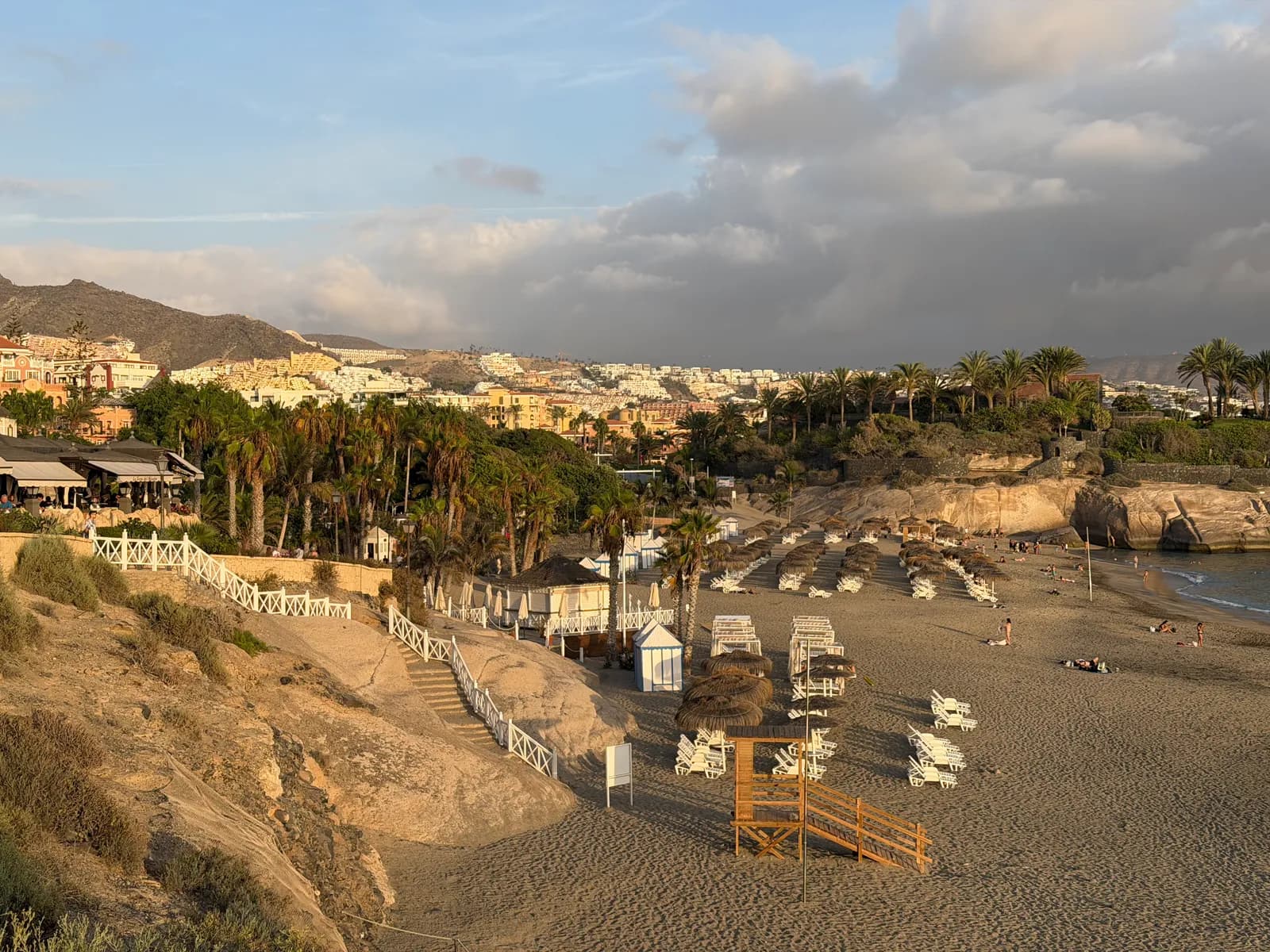 Playa del Duque w Costa Adeje wieczorem, drewniane zejście na plażę, parasole i zabudowa w tle