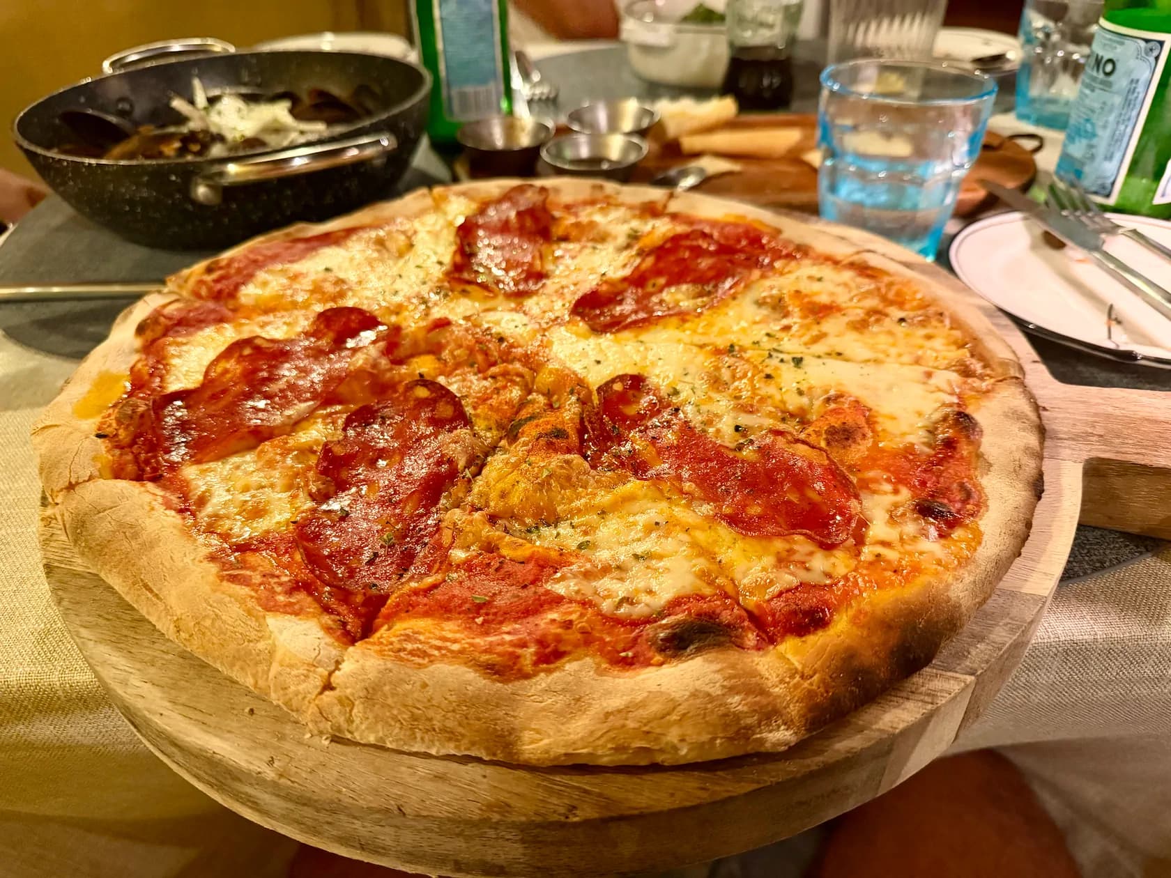Świeżo upieczona pizza z salami na drewnianej desce w restauracji, w tle kieliszki z wodą i oliwa