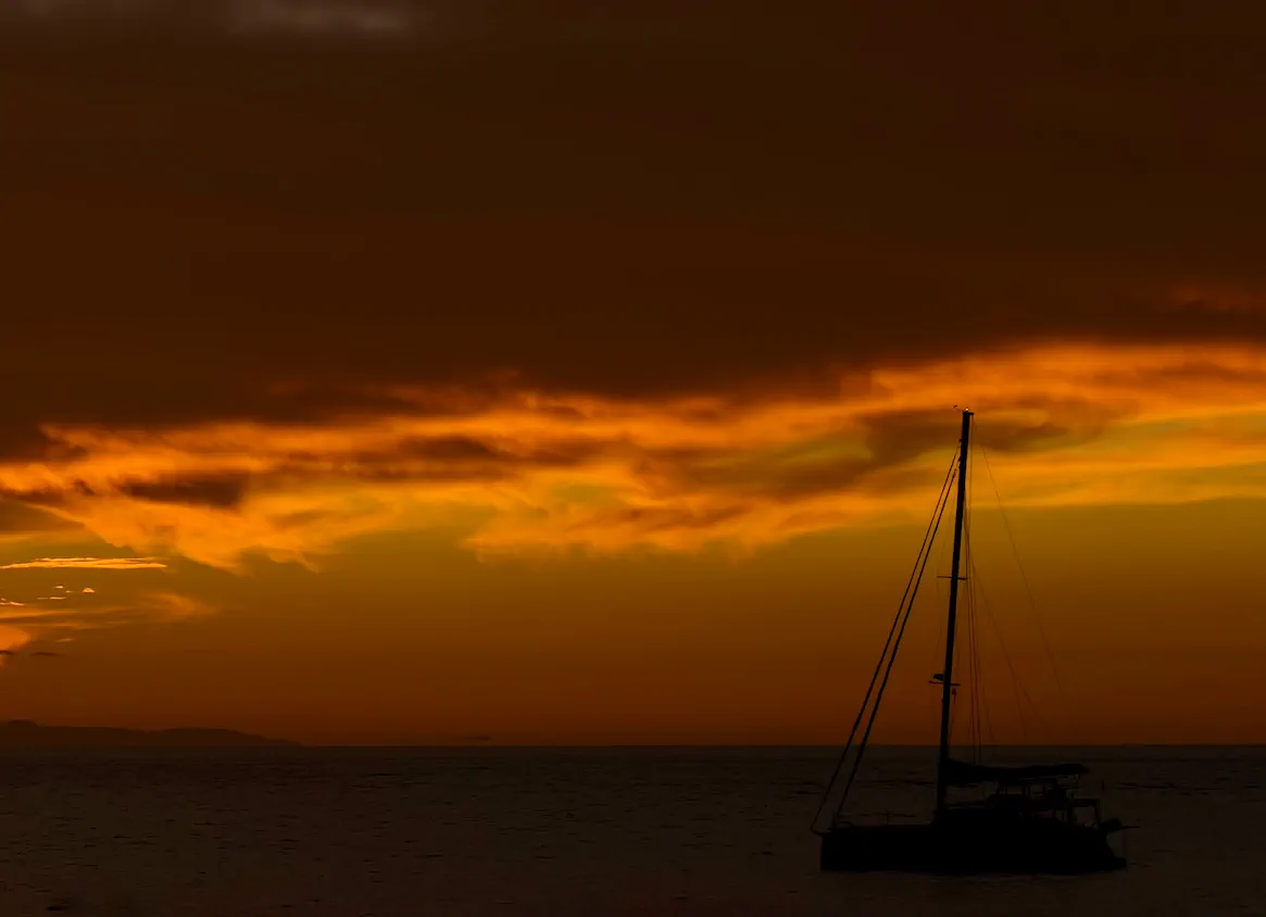 Sylwetka żaglówki na tle ciemnego oceanu i dramatycznego, pomarańczowo-złotego nieba podczas zachodu słońca