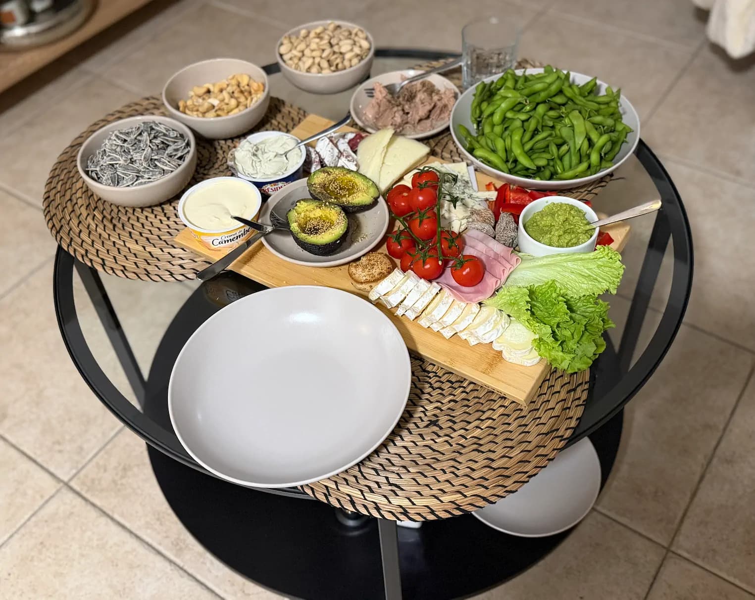 Szklany stół zastawiony zdrowymi przekąskami: awokado, pomidorki koktajlowe, edamame, sery, orzechy i nasiona