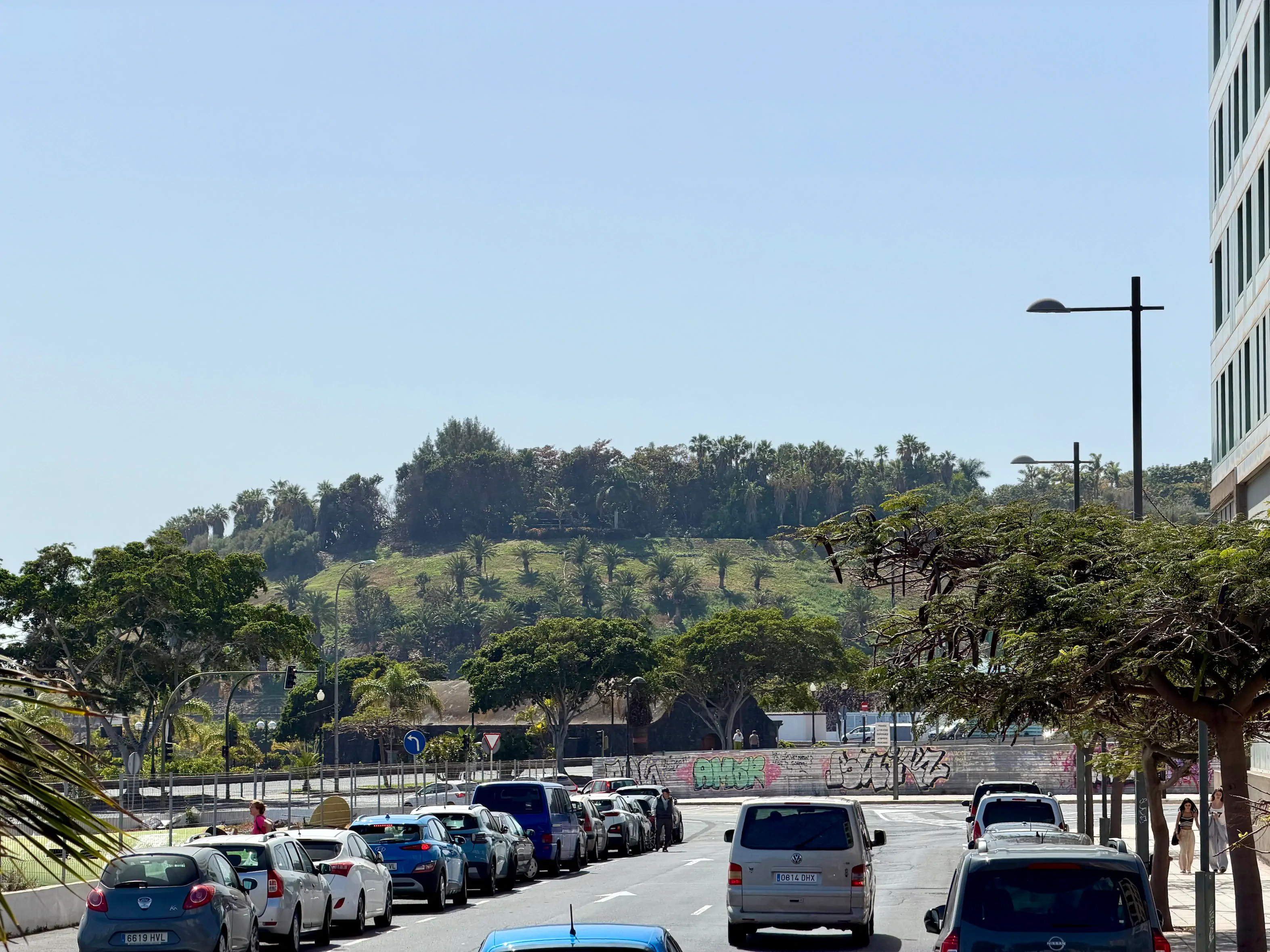 Słoneczna ulica na Teneryfie, zaparkowane samochody, a w tle zielone wzgórze porośnięte palmami